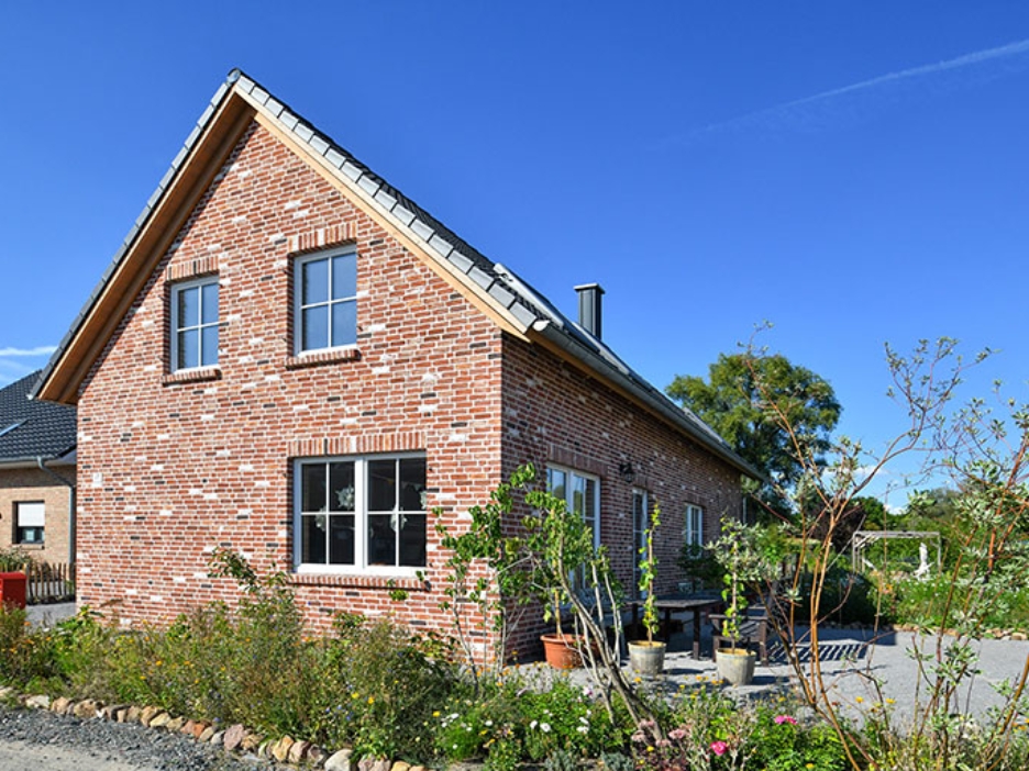 Gemütliches Backsteinhaus mit Satteldach, Garten und blühenden Pflanzen unter strahlend blauem Himmel.