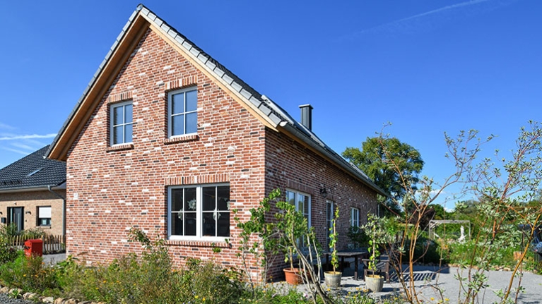 Gemütliches Backsteinhaus mit Satteldach, Garten und blühenden Pflanzen unter strahlend blauem Himmel.