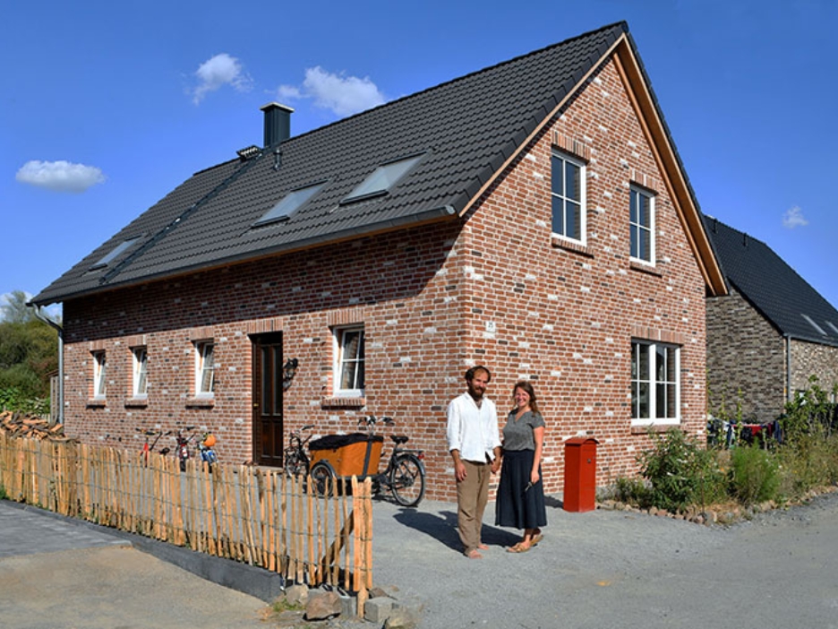 Backsteinhaus mit Satteldach und Garten, davor ein lächelndes Paar in der Sonne vor ihrem Zuhause.