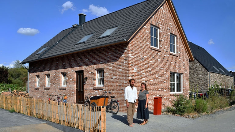 Backsteinhaus mit Satteldach und Garten, davor ein lächelndes Paar in der Sonne vor ihrem Zuhause.
