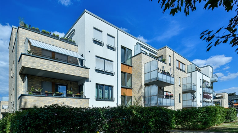 Helle Wohnhäuser mit Balkonen rahmen einen grünen Innenhof, in dem ein Spielplatz und schattige Bereiche Begegnungen ermöglichen.