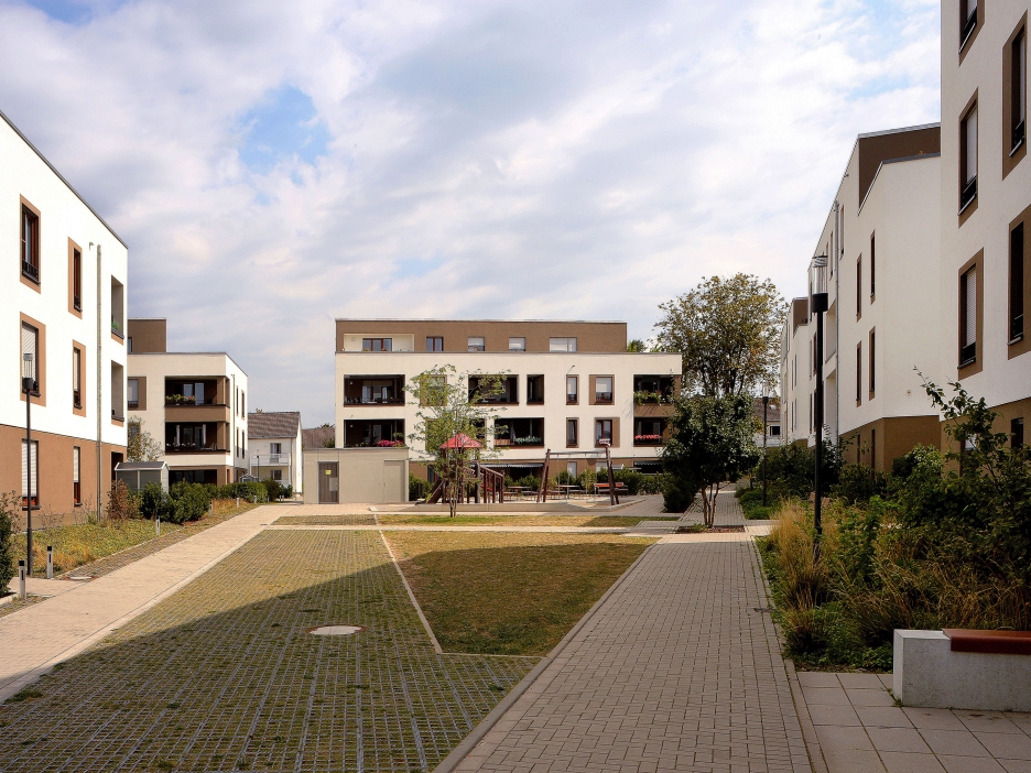 Weiße Wohngebäude formen einen großen Innenhof mit Spielbereich, Beeten und Wegen, der offen und freundlich gestaltet wirkt.
