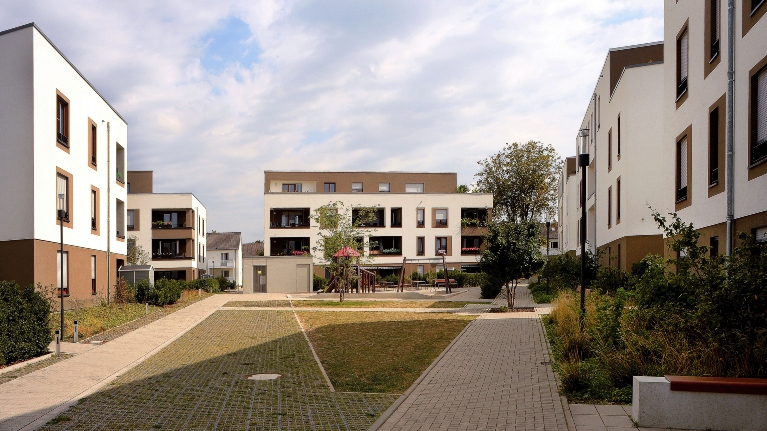 Weiße Wohngebäude formen einen großen Innenhof mit Spielbereich, Beeten und Wegen, der offen und freundlich gestaltet wirkt.