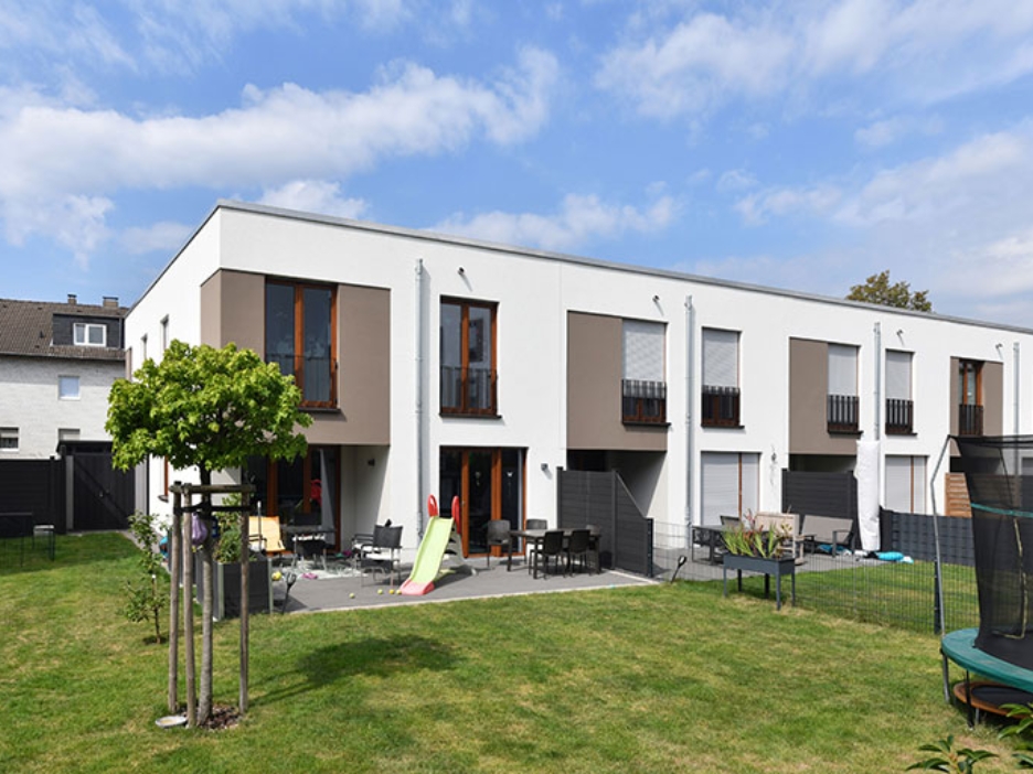 Moderne Reihenhäuser mit Terrassen, Spielgeräten und weitläufigem Garten unter blauem Himmel.
