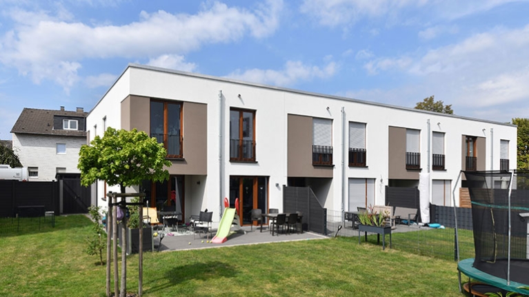 Moderne Reihenhäuser mit Terrassen, Spielgeräten und weitläufigem Garten unter blauem Himmel.