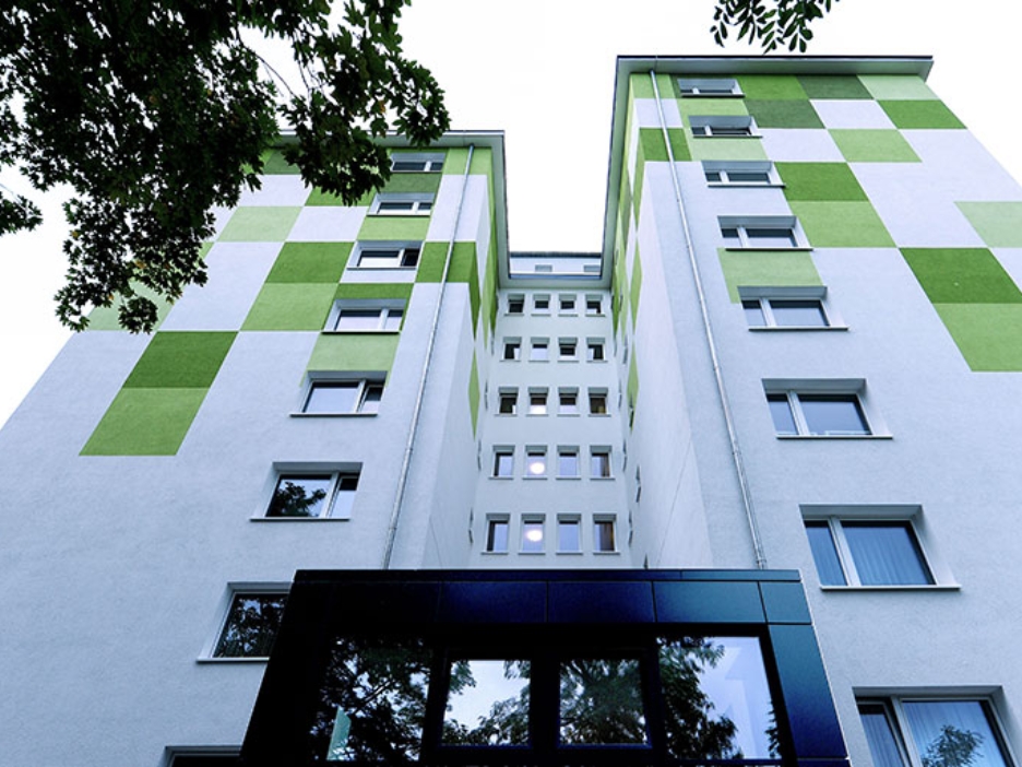 Hochhaus mit weißer Fassade vor blauem Himmel, flankiert von Bäumen am sonnigen Tag.