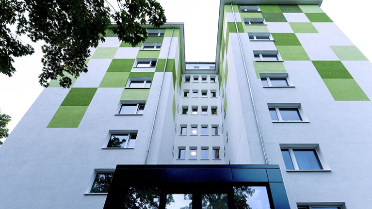 Hochhaus mit weißer Fassade vor blauem Himmel, flankiert von Bäumen am sonnigen Tag.