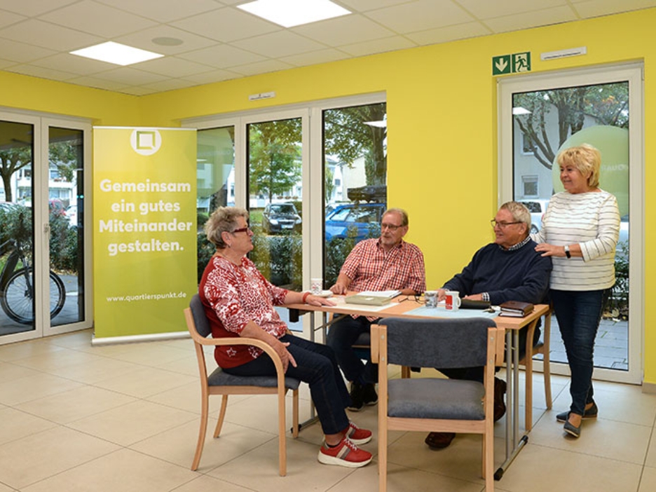 Eine kleine Gruppe trifft sich im hellen Raum vor gelber Wand, um sich auszutauschen und das nachbarschaftliche Miteinander zu stärken.