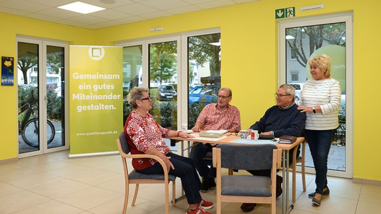 Eine kleine Gruppe trifft sich im hellen Raum vor gelber Wand, um sich auszutauschen und das nachbarschaftliche Miteinander zu stärken.