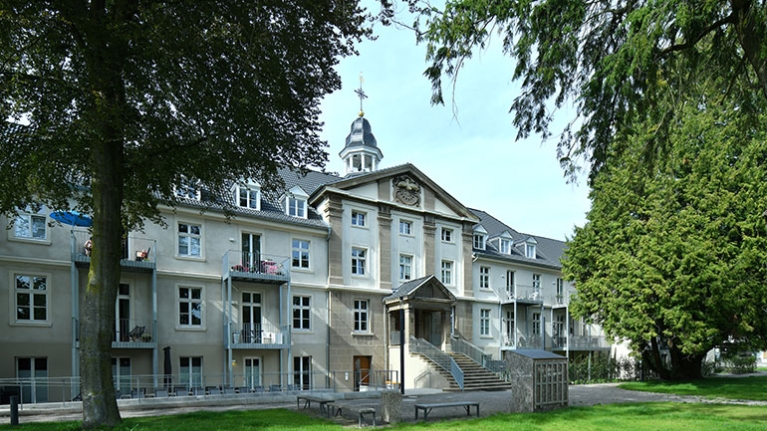 Historisches Klostergebäude mit Balkonen, großem Treppenportal und altem Baumbestand.