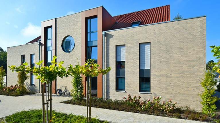 Ein modernes Gebäude mit hellem Klinker und rötlichem Dach steht zwischen jungen Bäumen und gepflegten Wegen im Quartier.