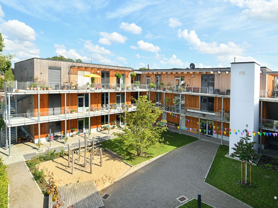 Offene Wohnanlage aus Holz mit Balkonen, Innenhof und großem Spielbereich bei sonnigem Wetter.