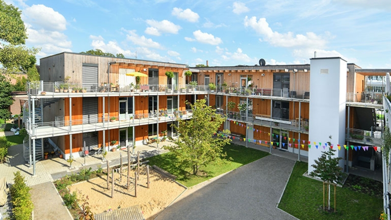 Offene Wohnanlage aus Holz mit Balkonen, Innenhof und großem Spielbereich bei sonnigem Wetter.