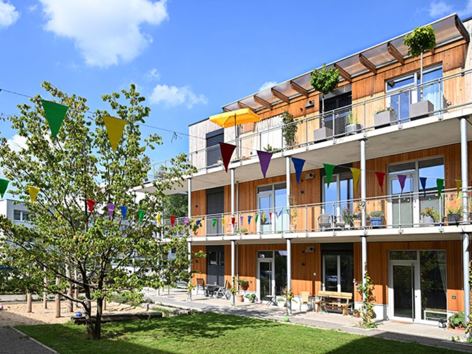 Holzbau mit bunten Wimpeln und grüner Mitte, Balkone voller Pflanzen unter blauem Himmel.