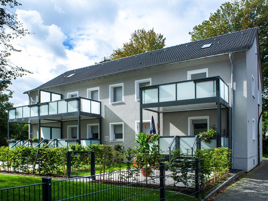 Graues Wohnhaus mit großen Balkonen und gepflegten Vorgärten unter leicht bewölktem Himmel.