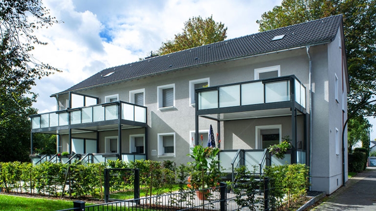 Graues Wohnhaus mit großen Balkonen und gepflegten Vorgärten unter leicht bewölktem Himmel.