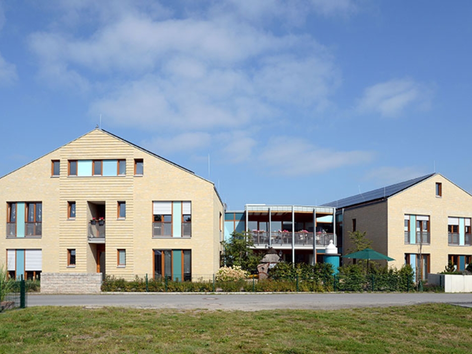 Zwei moderne Gebäude mit großen Fenstern bilden ein Ensemble, das mit Grün und offenen Bereichen freundlich in den Ort eingebettet ist.