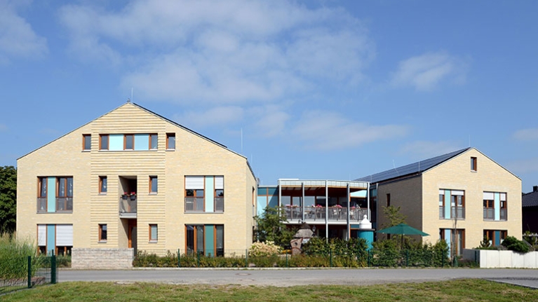 Zwei moderne Gebäude mit großen Fenstern bilden ein Ensemble, das mit Grün und offenen Bereichen freundlich in den Ort eingebettet ist.