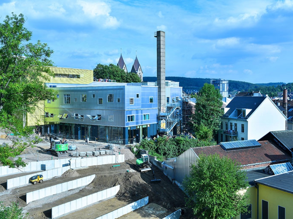 Silbern glänzender Neubau mit großem Schornstein, davor Terrassenbau und Baumaschinen.