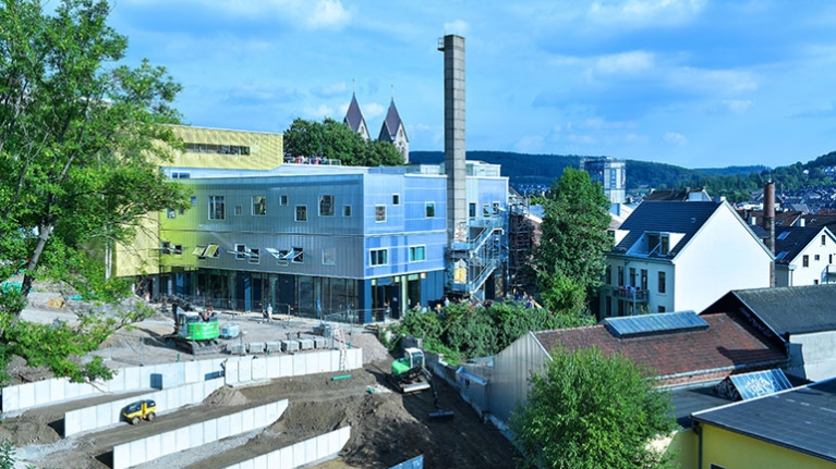 Silbern glänzender Neubau mit großem Schornstein, davor Terrassenbau und Baumaschinen.