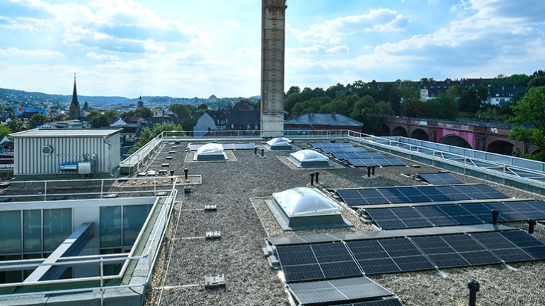 Auf dem Dach liegen Solarpaneele und Oberlichter, während sich dahinter die Stadt mit Kirchturm und grünen Hügeln ausbreitet.