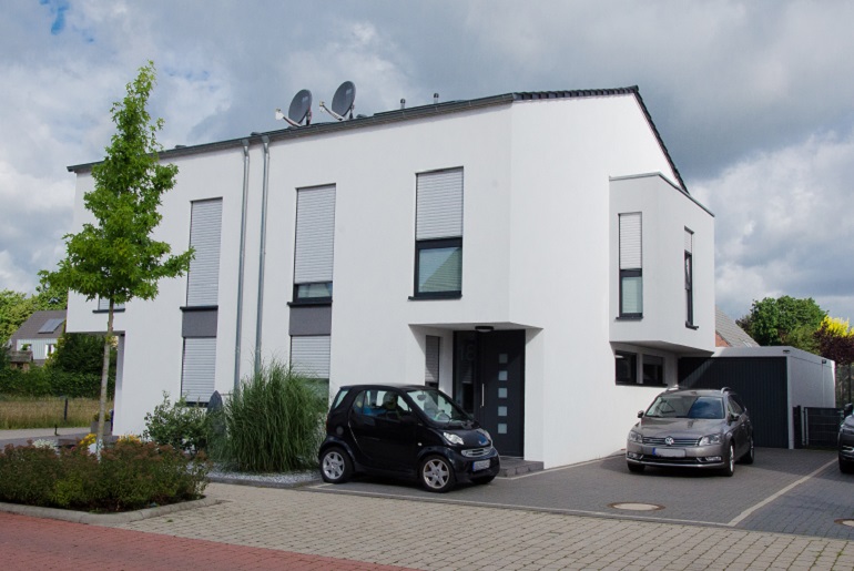 Eine weiß verputzte Doppelhaushälfte mit Garage und grau gepflasterter Einfahrt, auf der zwei Autos stehen.