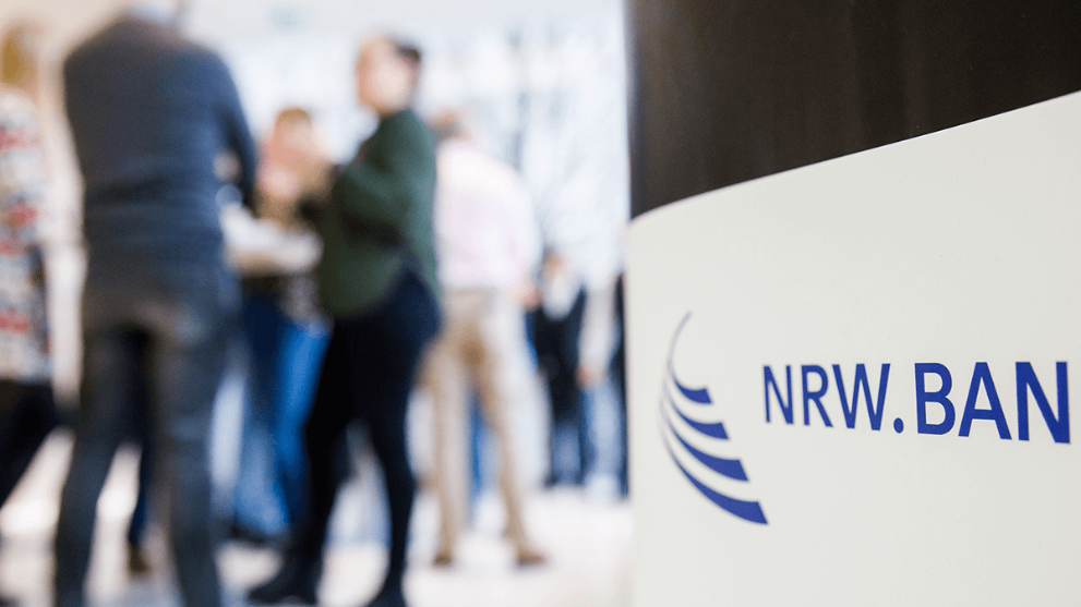 Mehrere Menschen unterhalten sich an Stehtischen. Im Vordergrund das NRW.BANK-Logo