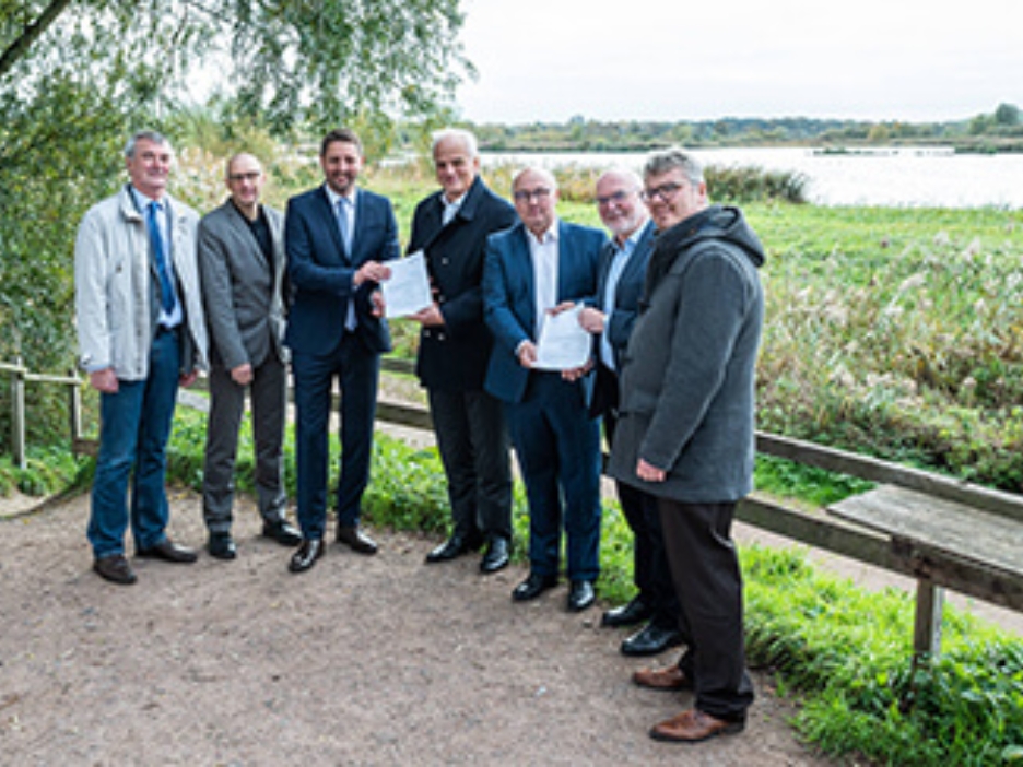 7 Männer bei der Übergabe eines Förderbescheids vor einer Seenlandschaft