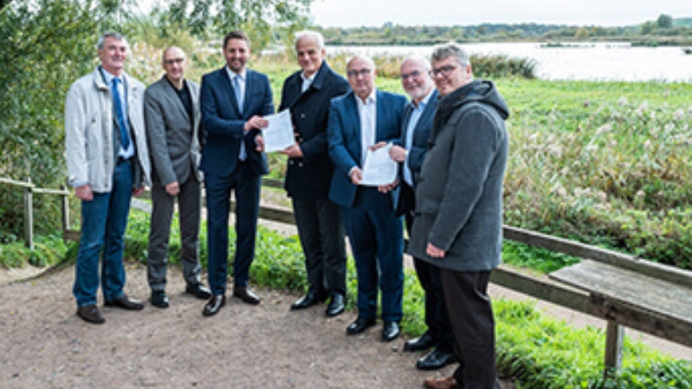 7 Männer bei der Übergabe eines Förderbescheids vor einer Seenlandschaft