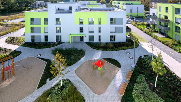 Grün-graue Häuser mit Flachdächern umrahmen einen begrünten Innenhof mit Spielplatz