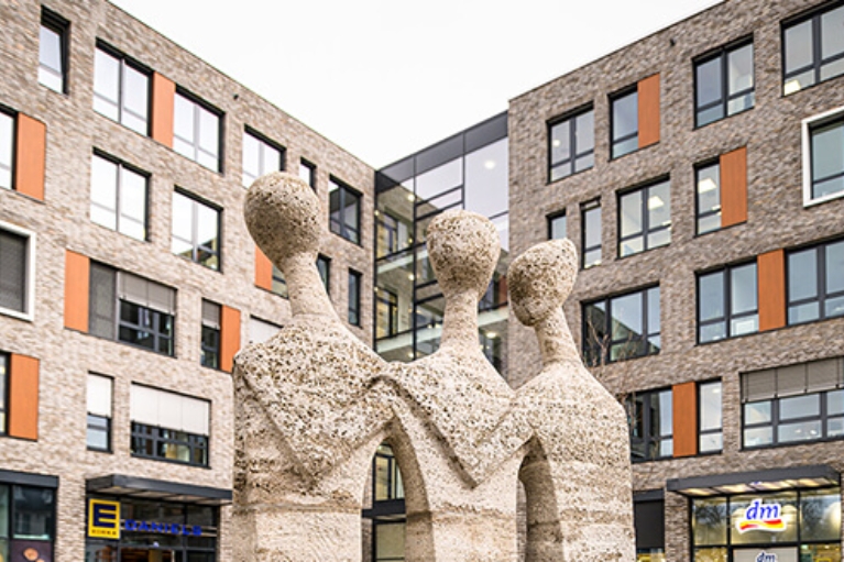 Steinernes Standbild vor einem modernen Gebäudekomplex mit Wohneinheiten, Super- und Drogeriemarkt