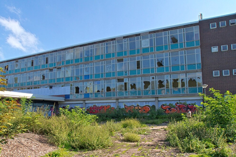 Außenansicht einer ehemaligen Schule mit zerstörten Fenstern, davor eine Brachfläche