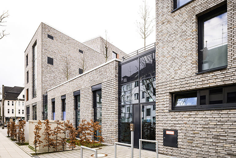 Ein Wohnhaus mit Flachdach und heller Klinkerfassade. Davor und darauf Bäume und Büsche mit herbstlichen Laub.
