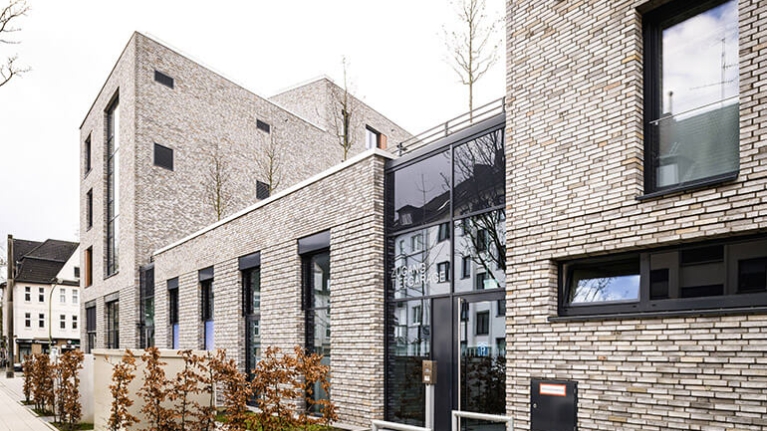 Ein Wohnhaus mit Flachdach und heller Klinkerfassade. Davor und darauf Bäume und Büsche mit herbstlichen Laub.
