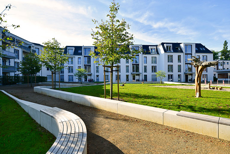 Ein Weg führt durch einen neu angelegten Park mit einer Wiese, einem Sandkasten und Sitzhelegenheiten. Im Hintergrund ist ein modernes Mehrfamilienhaus zu sehen.