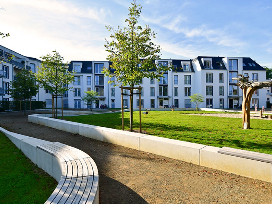 Ein Weg führt durch einen neu angelegten Park mit einer Wiese, einem Sandkasten und Sitzhelegenheiten. Im Hintergrund ist ein modernes Mehrfamilienhaus zu sehen.
