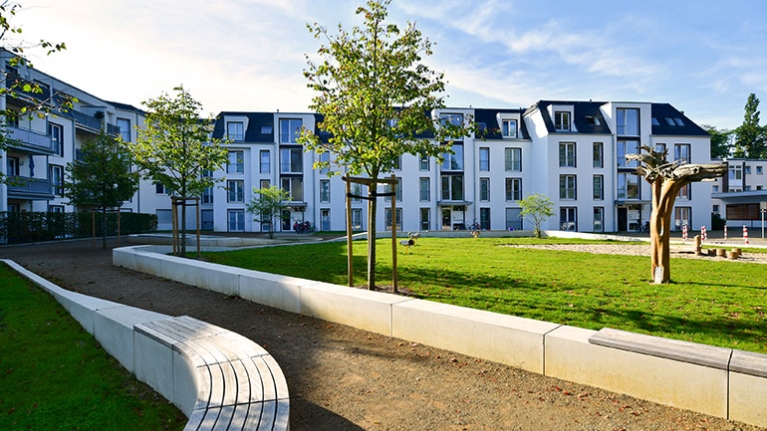 Ein Weg führt durch einen neu angelegten Park mit einer Wiese, einem Sandkasten und Sitzhelegenheiten. Im Hintergrund ist ein modernes Mehrfamilienhaus zu sehen.