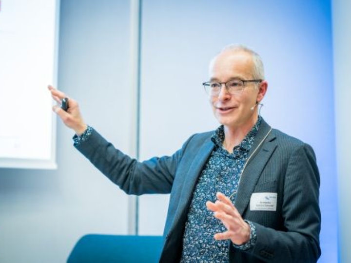 Ein Mann im Sakko steht vor einer Leinwand und erklärt etwas mit ausgestrecktem Arm und einem Presenter in der Hand.