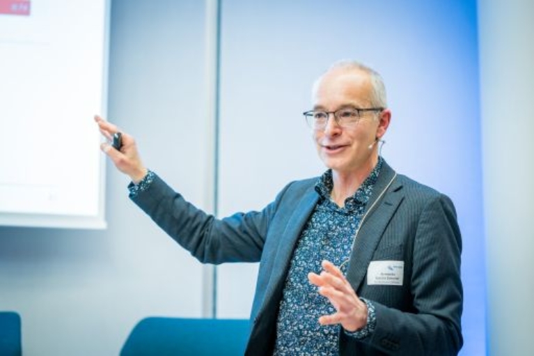 Ein Mann im Sakko steht vor einer Leinwand und erklärt etwas mit ausgestrecktem Arm und einem Presenter in der Hand.