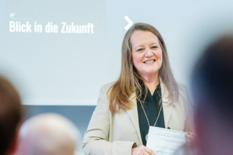 Eine Frau mit Mikrofon spricht vor Publikum, im Hintergrund ist der Schriftzug „Blick in die Zukunft“ zu sehen.