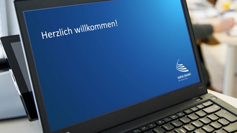 Auf dem Bildschirm eines Laptops steht mit weißer Schrift auf blauem Hintergrund: "Herzlich willkommen!"