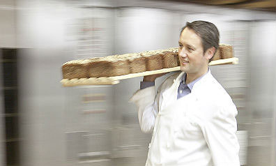 Ein Bäcker in weißem Kittel trägt ein Brett mit mehreren Broten