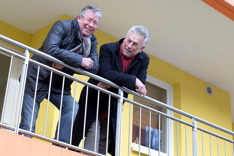 Zwei Männer beugen sich übers Geländer eines Balkons und lächeln in die Kamera