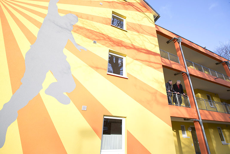 Ausschnitt der gelb-roten Fassade eines Hauses, zwei Männer stehen auf dem Balkon