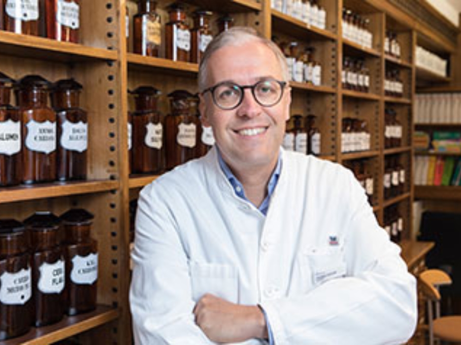 Ein Apotheker in weißem Kittel vor einem Regal mit Arzneiflaschen. Er lächelt in die Kamera.