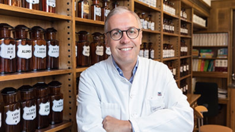 Ein Apotheker in weißem Kittel vor einem Regal mit Arzneiflaschen. Er lächelt in die Kamera.