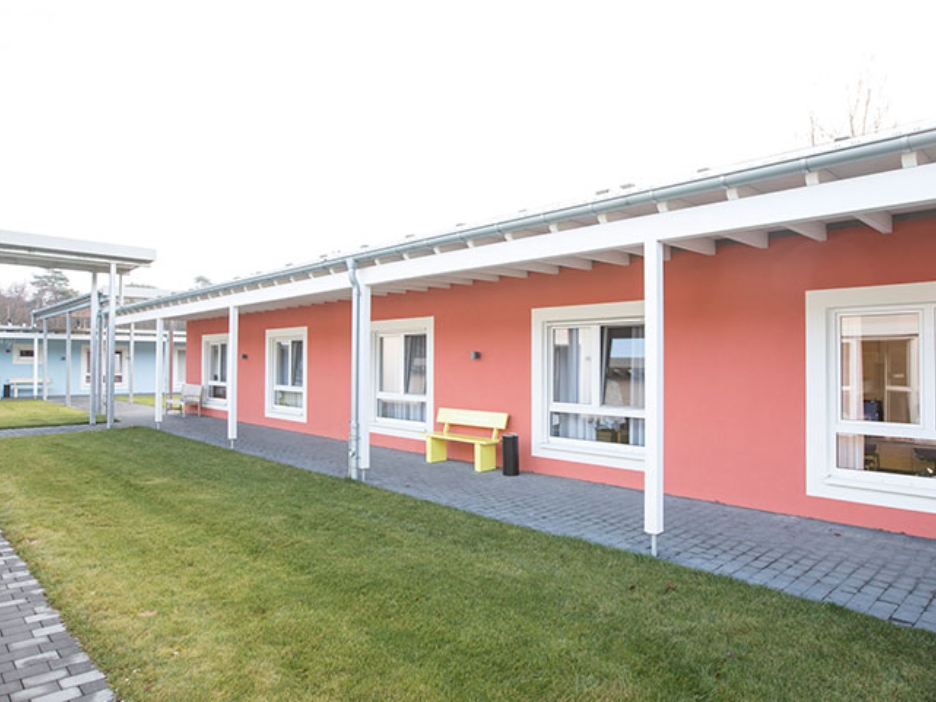 Rotes, eingeschossiges Gebäude, davor eine gelbe Bank, ein gepflasterter Weg und ein Grünstreifen