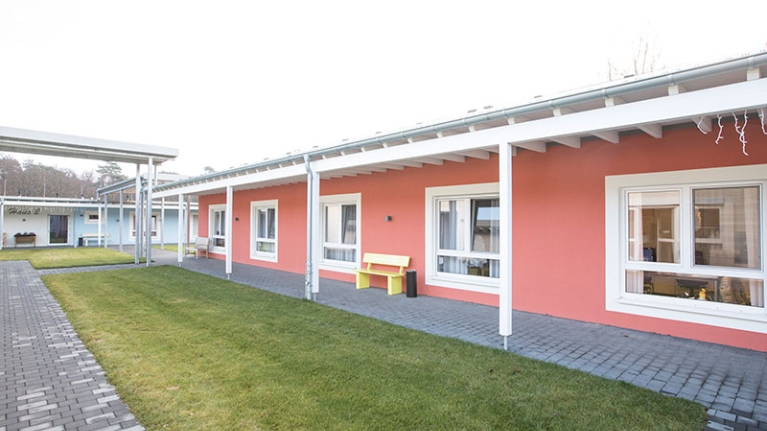 Rotes, eingeschossiges Gebäude, davor eine gelbe Bank, ein gepflasterter Weg und ein Grünstreifen