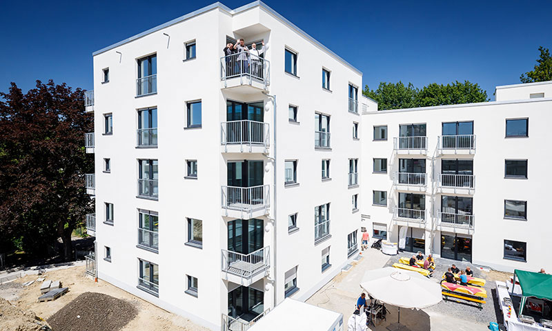 Modernes, weißes Mehrfamilienhaus , drei Menschen winken vom Balkon