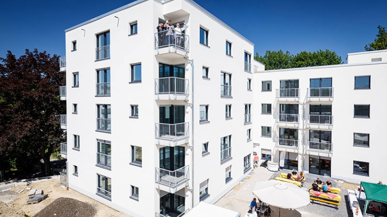 Modernes, weißes Mehrfamilienhaus , drei Menschen winken vom Balkon